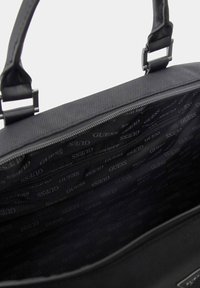 Borsa nera con una texture liscia, dotata di chiusura con zip e fodera interna a fantasia con il logo "GUESS". Inclusi manici regolabili.