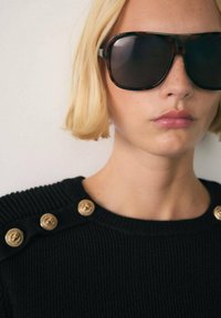 Occhiali da sole tartaruga con lenti scure, indossati da una persona in un maglione nero a coste con dettagli in bottone dorato sulle spalle.
