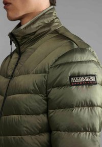 Giacca a vento verde con colletto alto, design trapuntato e zip nera; presenta una patch con il marchio sul braccio sinistro con accenti rossi.