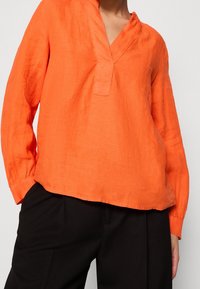Haut en lin orange à manches longues avec un col en V et un petit plastron à l'avant ; associé à un pantalon noir texturé, affichant une coupe décontractée et un design casual.