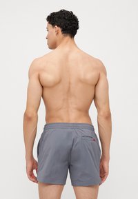 Shorts de bain gris avec une taille élastique, poches latérales et une seule poche arrière avec un petit logo rouge. Tissu lisse et léger.