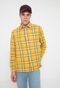 Gelbes kariertes Hemd mit blauen und braunen Akzenten, zwei Brusttaschen und Knopfverschluss. Getragen mit hellblauen Jeans und einer burgunderfarbenen Kappe.