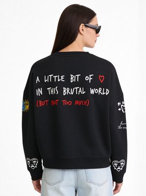 Femme portant des lunettes de soleil, vêtue d’un sweat noir avec des patchs colorés sur les manches et un texte audacieux inscrit dans le dos : « A LITTLE BIT OF LOVE IN THIS BRUTAL WORLD (BUT NOT TOO MUCH). »