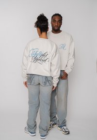 Twee personen dragen bijpassende witte sweaters en lichtblauwe spijkerbroeken, één kijkt naar voren, de ander toont de tekst "Multiply Apparel" op de achterkant.