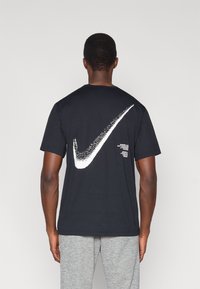 Μαύρο βαμβακερό t-shirt με μεγάλο λευκό γραφικό Nike swoosh στην πλάτη, με επιπλέον κείμενο κάτω από το σχέδιο.