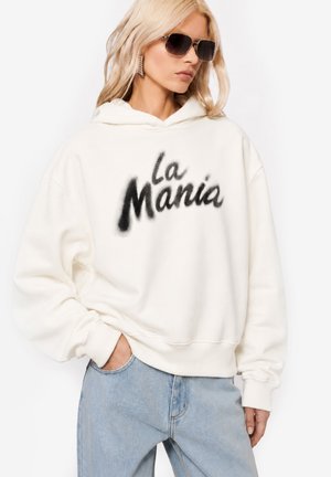 Kobieta w okularach przeciwsłonecznych, białej bluzie z kapturem z napisem "La Mania" czarnymi literami, jasnoniebieskich dżinsach i z jedną ręką w kieszeni.