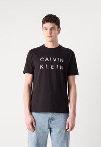Tricou negru din bumbac cu guler rotund, având litere mari, metalice, de culoare argintie "CALVIN KLEIN" pe față. Purtați cu blugi decolorați.