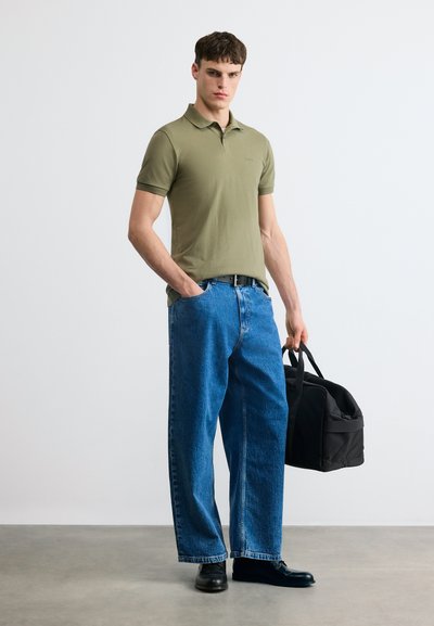 Jeune homme portant un polo vert olive, un jean bleu et des chaussures noires, tenant un sac de sport noir dans une main, debout devant un fond uni.