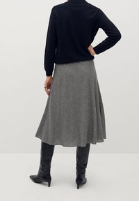 Mujer vestida con una blusa negra de manga larga, una falda midi de patrón gris y botas altas de tacón negro, de pie con una mano en la cadera.