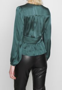 Blouse en satin vert émeraude avec manches bouffantes, taille cintrée et détail plissé au dos, associée à un pantalon en cuir noir.
