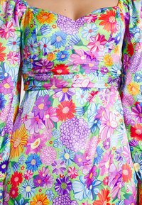 Kukkapaikkakuviolla varustettu mekko, jossa on monia värejä vihreällä taustalla, puffihihat ja vartalonmyötäinen yläosa. Huomaa näkyvät pleattytykset.