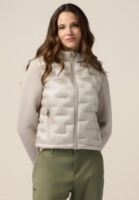 Vest ușor bej umplut, cu un tipar structurat, guler înalt, închidere cu fermoar și buzunare laterale. Purta peste un top cu mânecă lungă și pantaloni verzi.
