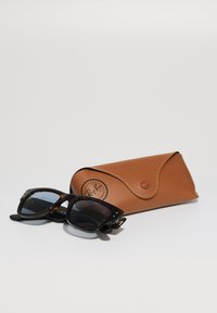Ochelari de soare din tortoiseshell negru cu rame rectangulare, însoțiți de un etui din piele texturată maronie, cu un logo.