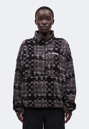 Person iført en sort og grå ternet fleece-pullover med halv lynlås og Columbia-logo på brystet, stående mod en ensfarvet baggrund.