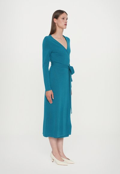 Tealgrön omlottklänning med långärmade ärmar, tillverkad av texturerat tyg, med skärp som knyts i midjan och en midilång kjol. Par av off-white klackskor.