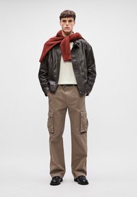 Pruun nahktagi, kreemjas kampsun, rustikarbne sall, beezid cargo-püksid taskutega ja mustad kingad. Modell seisab ühtlase tausta ees.