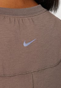 Camiseta deportiva marrón con textura de canalé, que presenta un pequeño logo de Nike en azul claro en la parte posterior cerca del escote. Detalle de costura plana visible.
