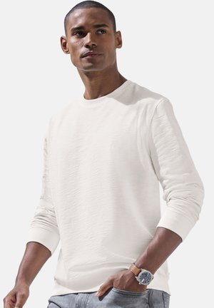 Man wearing white long-sleeve shirt and blue jeans, with wristwatch, looking away against a plain light background.