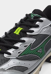 Pantof sport negru și gri din plasă, cu suprapuneri argintii, logo Mizuno verde, șireturi negre și o etichetă mică galben neon pe limbă.