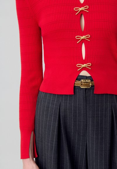 Maglione rosso a coste con tre accenti a fiocco dorato, caratterizzato da ritagli e spaccature laterali. Abbinato a pantaloni grigi a righe sottili e una cintura color marrone chiaro.