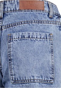 Pantalones vaqueros de mezclilla azul claro con un bolsillo trasero y un parche rectangular de cuero marrón en la cinturilla, mostrando costuras y textura cosidas.