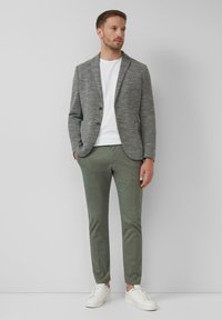 Mann in einem grauen strukturierten Blazer, einem weißen T-Shirt, olivgrünen Hosen und weißen Turnschuhen, der vor einer schlichten hellgrauen Wand steht.
