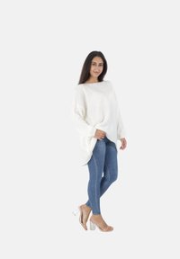 Witte oversized trui met een wijd decolleté, gecombineerd met strakke blauwe spijkerbroek en transparante haksandalen. Over het geheel een gladde textuur.