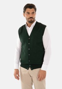 Gilet lavorato a maglia verde scuro con scollatura a V, dotato di sei bottoni frontali, indossato sopra una camicia bianca con colletto, abbinato a pantaloni beige.