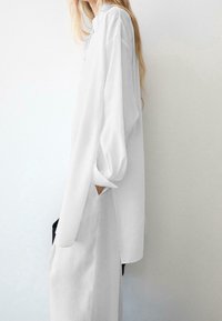 Personne aux longs cheveux blonds porte une robe chemise blanche ample avec la main dans la poche latérale, debout devant un fond clair.