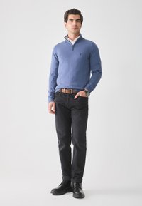 Mann som står i blå kvart-zip genser, hvit skjorte med krage, svarte jeans med brun belte, svarte støvler og klokke mot en ensfarget bakgrunn.