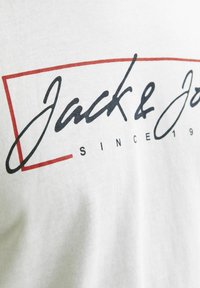 Hvid bomulds T-shirt med trykt tekst i sort og rød. Designet inkluderer "Jack & Jones" og "SINCE 1990" inden for en rektangulær ramme.