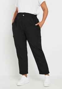 Pantalones negros de corte relajado con cintura elástica, bolsillos delanteros y puños doblados, combinados con zapatillas blancas para un look informal.