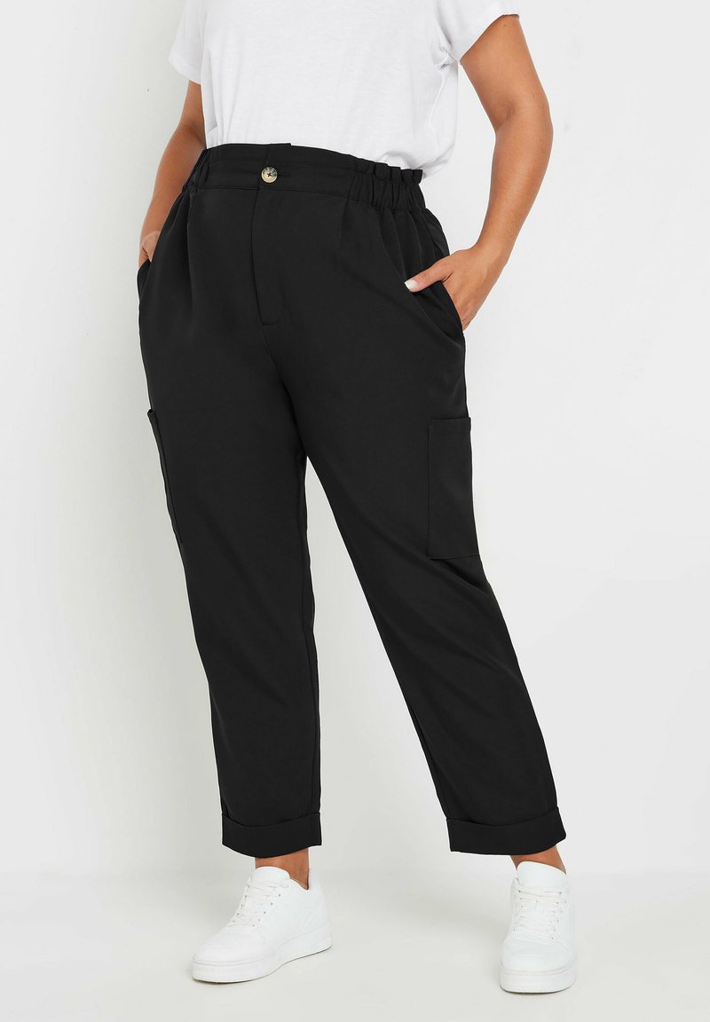 Pantalones negros de corte relajado con cintura elástica, bolsillos delanteros y puños doblados, combinados con zapatillas blancas para un look informal.