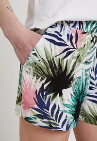 Shorts colorés en tissu avec imprimé de feuilles tropicales dans des nuances de vert, rose et bleu ; comprennent des poches latérales et une ceinture élastique.