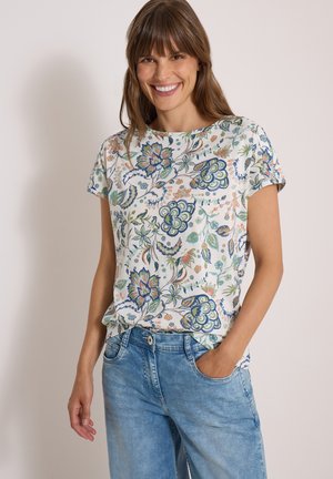 Femme souriante aux cheveux bruns portant un chemisier blanc à fleurs à manches courtes et un jean bleu clair, une main dans la poche.