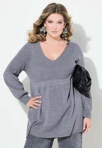 Pull gris à côtes avec un décolleté en V et des manches longues, présentant une coupe ample et des fentes sur les côtés, associé à une pochette en cuir noir.