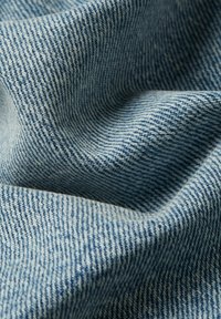 Jasnoniebieska tkanina dżinsowa z teksturowanym, prążkowanym wzorem. Materiał jest miękki i lekko pomarszczony, ukazując różne odcienie niebieskiego.