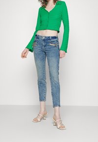 Top corto texturizado verde con escote en V y cierre de botones, combinado con jeans ajustados de color azul claro con bolsillos con cremallera y sandalias de tacón beige.