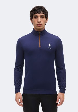 Homme portant un pull bleu marine à manches longues avec une fermeture éclair au quart et un logo blanc sur la poitrine gauche, debout devant un fond clair uni.