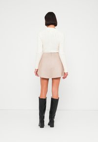 Κρεμ πουλόβερ με πλεκτό σχέδιο καλωδίων, μπεζ μίνι φούστα από σουέτ και μαύρα μπότες με τακούνι μέχρι το γόνατο. Λεπτή υφή και εφαρμοστό σχέδιο, πίσω όψη.