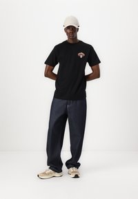 Schwarzes T-Shirt aus Baumwolle mit kurzen Ärmeln und einem bunten Grafiklogo auf der Brust, kombiniert mit locker sitzenden dunkelblauen Jeans und beigen Turnschuhen.