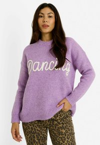 Mujer vistiendo un suéter de punto lila con el texto "Dancing" y pantalones de leopardo, posando con una mano en el bolsillo sobre un fondo blanco.