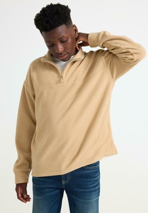 Beige fleece-paita, jossa on puolivetoketju, pehmeä rakenne, rento malli ja pitkät hihat. Vaatetta on käytetty vaalean värisen paidan päällä ja yhdistetty farkkuihin.