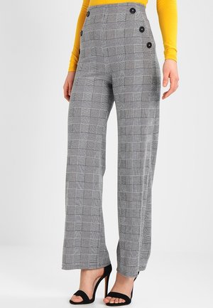 Nainen pukeutuneena harmaisiin ruudullisiin korkeavyötäröisiin housuihin, joissa on sivunapit, ja mustiin korkokenkiin, yhdistetty keltaiseen pitkähihainen paitaan.