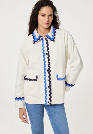 Frau trägt eine Cremejacke mit blau-schwarzem, wellenförmigem Besatz am Kragen, vorne, an den Taschen und Bündchen, kombiniert mit blauen Jeans.