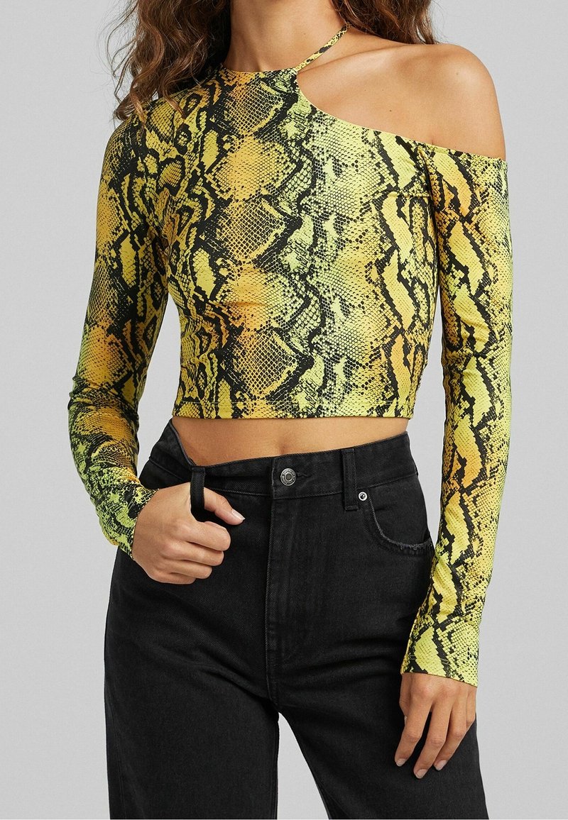 Långärmad croptop med en axel design, med mönster av en grön och gul orm. Bärs med svarta högmidjade jeans.