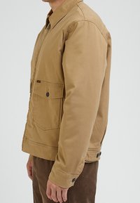 Beige Baumwolljacke mit durchgehender Knopfleiste, Brusttasche und seitlichen Taschen. Verfügt über einen Kragen und eine glatte, strapazierfähige Textur.