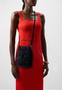 Czarna nylonowa torba typu worek z zapięciem na sznurek i przednią kieszenią na zamek, połączona z dopasowaną czerwoną sukienką z grubymi ramiączkami i teksturowanym materiałem.