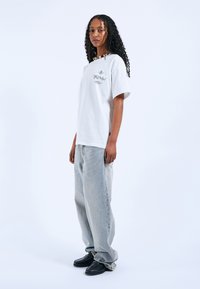 Bílý bavlněný tričko s grafickým potiskem, spárované s volnými světlými modrými džíny a černými botami, s hladkou texturou a uvolněným střihem.