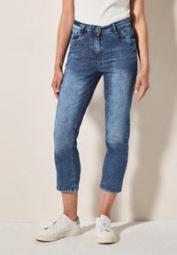 Jean en denim avec une finition bleue délavée, taille mi-haute, longueur cropped et design classique à cinq poches, associés à des baskets blanches.